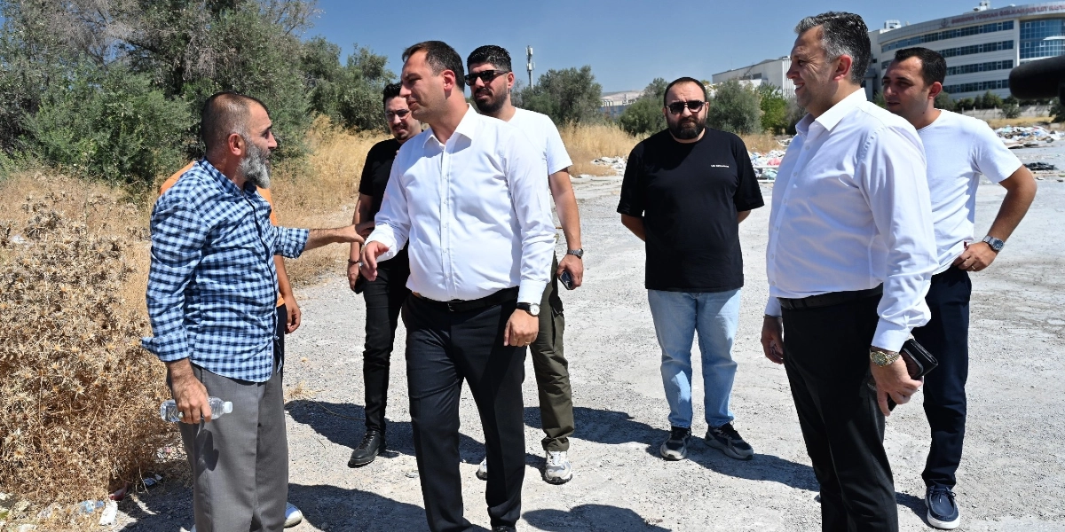 Açık havada, moloz dökülmüş boş bir arazide bir araya gelen bir grup erkek, çevredeki durumu inceliyor. Beyaz gömlekli ve siyah pantolonlu iki yetkili, yanlarındaki diğer kişilerle birlikte konuşarak alanı denetliyor. Arka planda kurumuş bitki örtüsü ve uzakta bir hastane binası görülüyor.