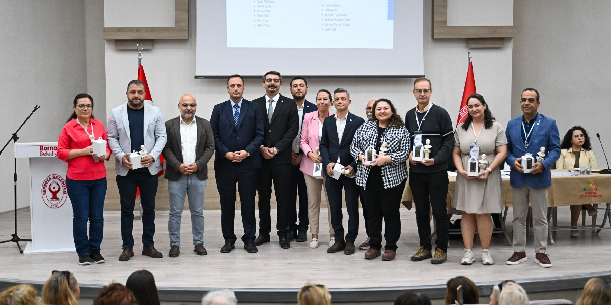 Bornova Belediyesi'nde düzenlenen bir etkinlikte, ellerinde küçük hediye kutuları ve büstler tutan, resmi kıyafetli bir grup insan sahnede yan yana durarak poz veriyor. Arka planda Bornova Belediyesi logosunun bulunduğu bir kürsü, Türk bayrakları ve üzerinde isim listesinin yer aldığı bir projeksiyon perdesi bulunuyor. En sağdaki masada ise Bornova Kent Konseyi logosu yer alıyor.