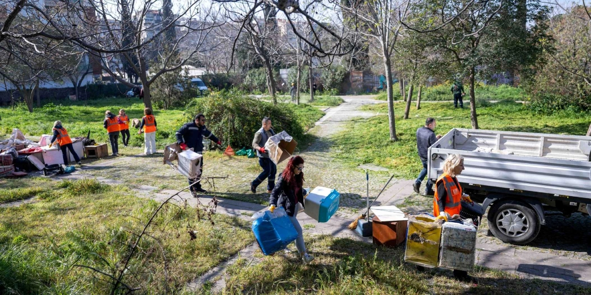 Turuncu yelek giyen bir grup gönüllü, Kedili Park'ta temizlik ve düzenleme çalışması yapıyor. Bazı kişiler ellerinde kedi evleri ve taşıma kutularıyla parkın içinde yürürken, bazıları da birikmiş atıkları topluyor. Sağ tarafta park edilmiş gri bir kamyonet ve çevrede ağaçlıklı yeşil bir alan görülüyor.