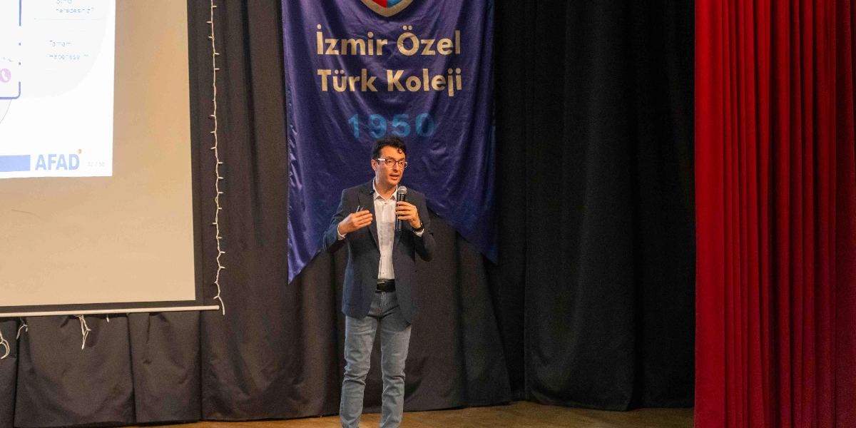 Koyu renkli bir ceket ve açık renkli pantolon giyen, gözlüklü bir erkek konuşmacı, sahnede elinde mikrofonla sunum yapıyor. Konuşmacının arkasında üzerinde 