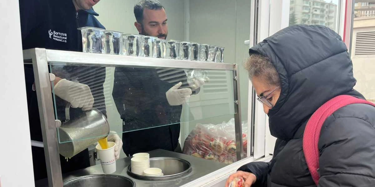 Bornova Belediyesi personeli, üzerinde kurum logosu bulunan kıyafetleri ve eldivenleriyle bir stantta, sıra bekleyen bir vatandaşa sıcak içecek ikram ediyor. Tezgahın üzerinde dizili boş karton bardaklar ve paketlenmiş yiyecekler bulunuyor.