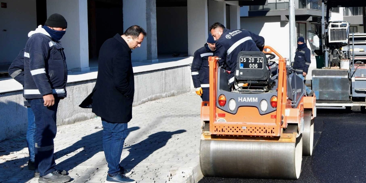 Bornova Belediyesi ekiplerinin yol yapım çalışması yürüttüğü alanda, siyah paltolu bir yetkili asfalt silindirini ve devam eden çalışmaları inceliyor. İş kıyafetli belediye personeli, üzerinde Bornova Belediyesi logosu bulunan turuncu renkli Hamm marka silindirin başında görev yapıyor.