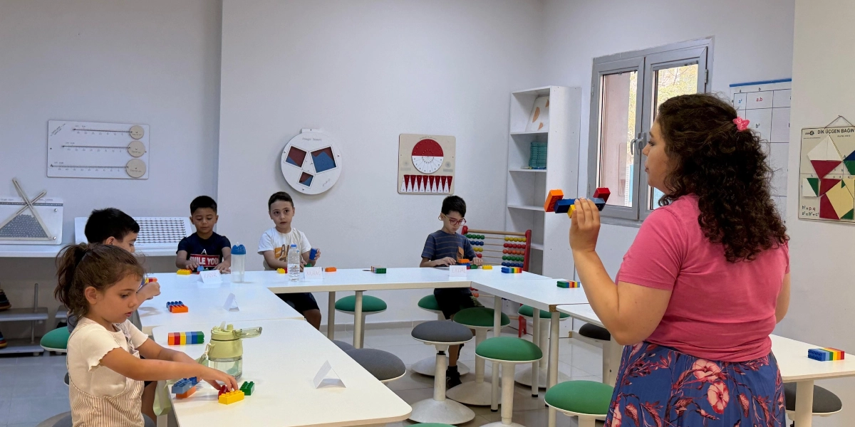 Aydınlık bir sınıfta, U şeklindeki masaların etrafında oturan beş çocuk ve onlara rehberlik eden pembe tişörtlü bir eğitmen renkli plastik tuğlalarla etkinlik yapıyor. Eğitmen elinde tuttuğu birleştirilmiş tuğlaları gösterirken, çocuklar önlerindeki masalarda kendi tuğlalarıyla çalışıyor. Duvarlarda matematiksel kavramları ve geometrik şekilleri gösteren eğitici panolar asılı bulunuyor.