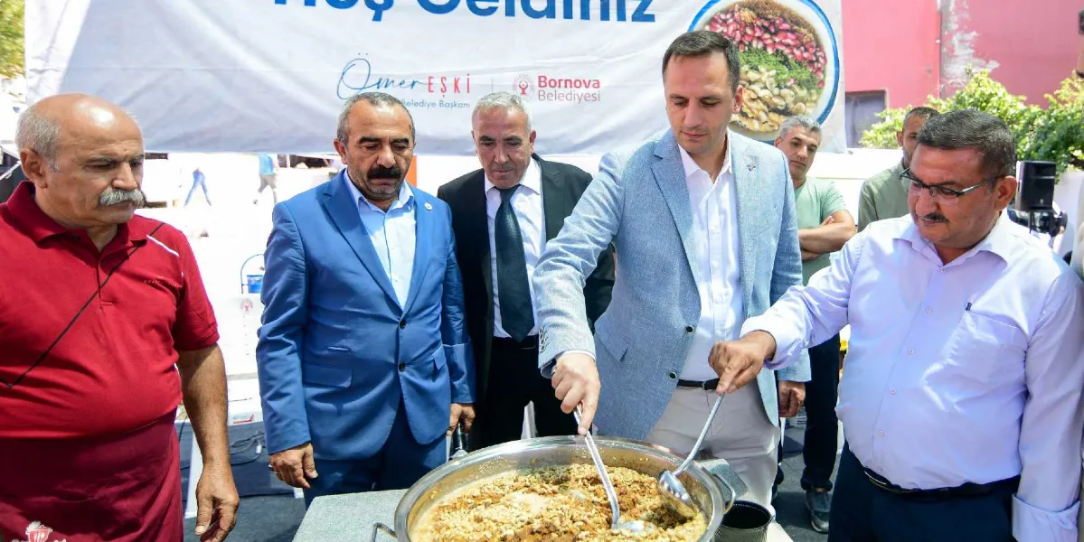 Bornova Belediye Başkanı Ömer Eşki ve beraberindeki heyet, büyük bir kazanda hazırlanan aşureyi kepçeyle karıştırarak vatandaşlara dağıtıyor. Arka planda üzerinde 