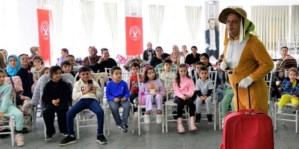Aydınlık bir salonda, üzerinde sarı tüylü bir kostüm ve geniş kenarlı bir şapka bulunan tiyatro oyuncusu, elinde kırmızı bir bavulla çocuklara gösteri yapıyor. Oyuncuyu ilgiyle izleyen çocuklar ve onlara eşlik eden yetişkinler beyaz sandalyelerde oturuyor. Arka planda Bornova Belediyesi logolu kırmızı flamalar ve Mustafa Kemal Atatürk’ün bir posteri yer alıyor.