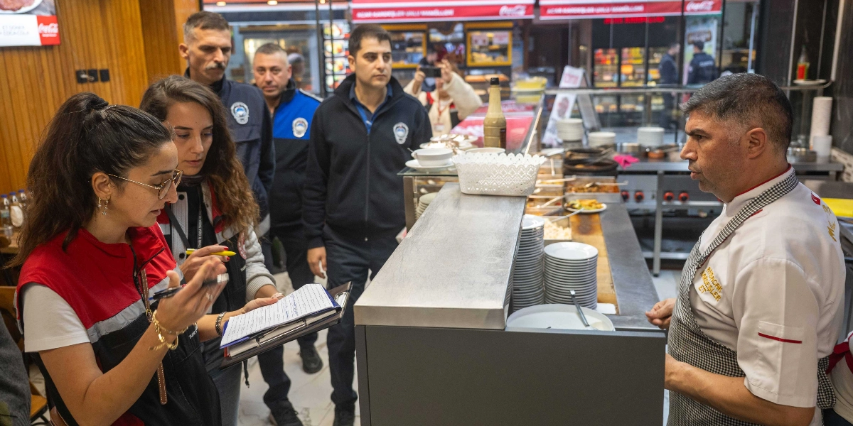 Zabıta ekipleri ve gıda denetçileri, bir restoranın mutfak bölümünde beyaz önlüklü bir aşçı ile görüşerek notlar alıyor. Denetim yapan görevliler ellerindeki evrakları incelerken, aşçı tezgahın arkasında durarak onlara yanıt veriyor. Arka planda restoranın servis alanı ve mutfak ekipmanları görülüyor.