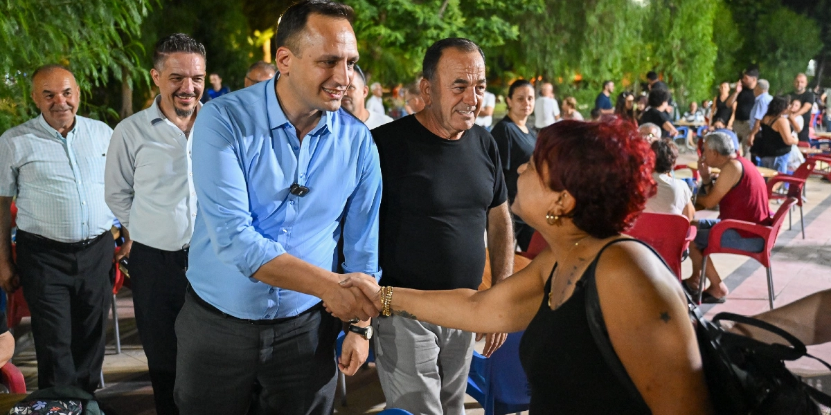 Mavi gömlekli bir erkek, açık havada düzenlenen bir etkinlikte kırmızı saçlı bir kadınla el sıkışıyor. Arka planda siyah tişörtlü bir adam ve diğer katılımcılar yer alırken, ortamda ağaçlar ve masalar bulunuyor.