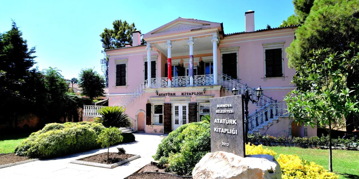 Bornova Belediyesi Atatürk Kitaplığı'nın dış cephesi. Pembe renkli, sütunlu ve balkonlu tarihi binanın önünde, üzerinde 