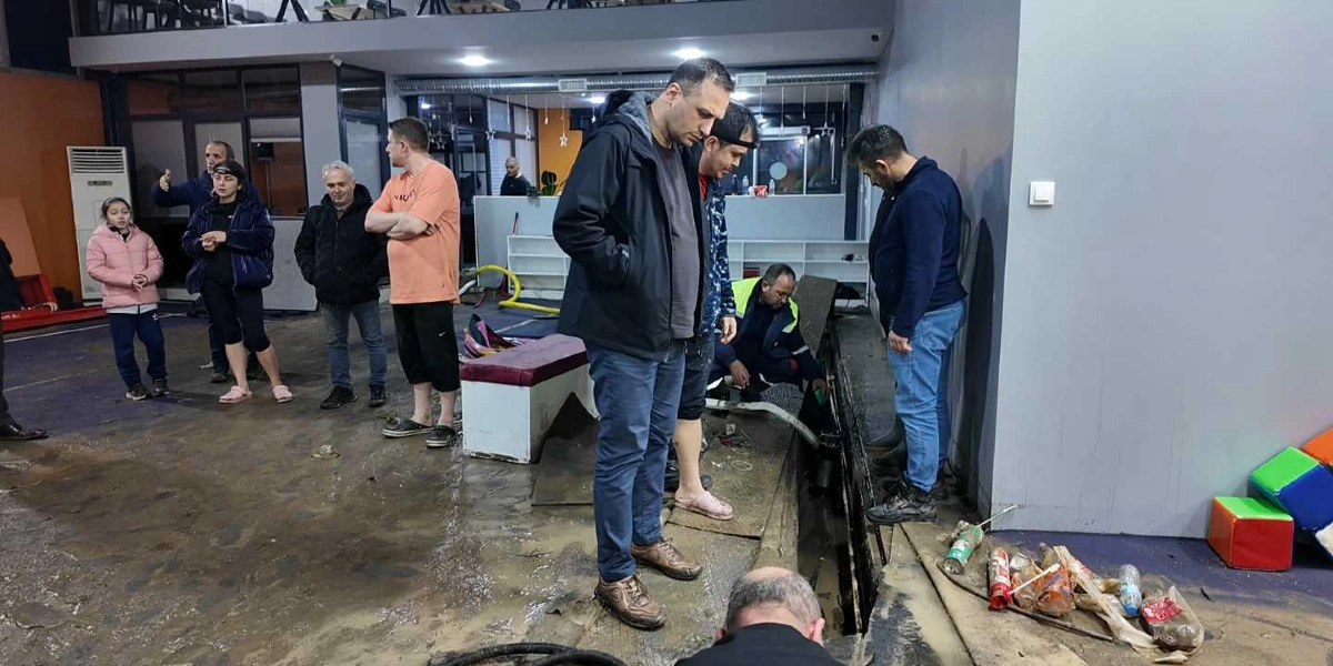 İç mekanın zeminini kaplayan çamurlu su birikintisi içerisinde, bir grup insan selin etkilerini incelemektedir. Bir görevli, zemindeki bir boşluğun içine hortum yerleştirerek su tahliyesi yapmaya çalışırken, çevresindeki yetişkinler ve bir çocuk endişeli bir şekilde süreci izlemektedir. Ortamda dağınık halde duran plastik şişeler ve çocuk oyun alanına ait renkli küpler dikkat çekmektedir.