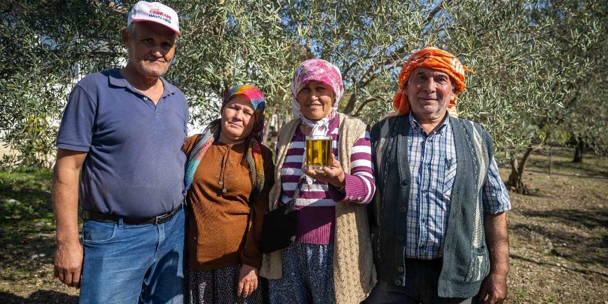 Zeytin ağaçlarının önünde yan yana duran, başlarında yöresel eşarplar bulunan iki kadın ve iki erkek, gülümseyerek kameraya bakıyor. Kadınlardan biri elinde bir bardak dolusu zeytinyağı tutuyor.