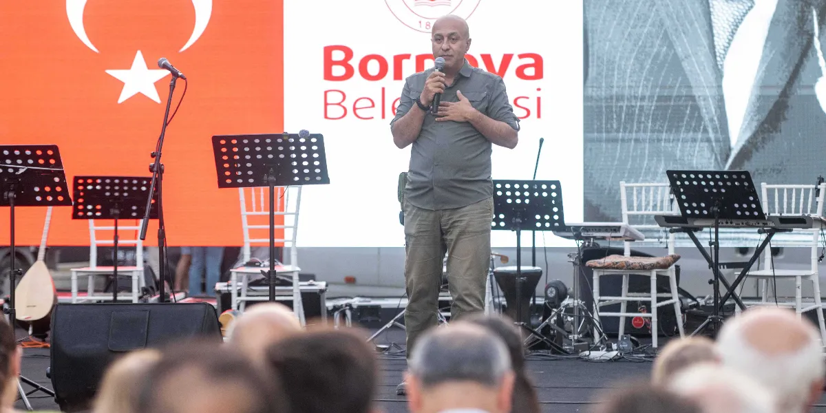 Gri gömlekli ve haki pantolonlu orta yaşlı bir erkek, elinde mikrofonla sahnede konuşma yapıyor. Bir elini kalbinin üzerine koymuş olan konuşmacının arkasında, üzerinde Türk bayrağı ve Bornova Belediyesi logosunun yer aldığı büyük bir ekran bulunuyor. Sahnede ayrıca müzik aletleri, nota sehpaları ve beyaz sandalyeler yer alıyor; ön planda ise izleyicilerin başlarının silüetleri görülüyor.