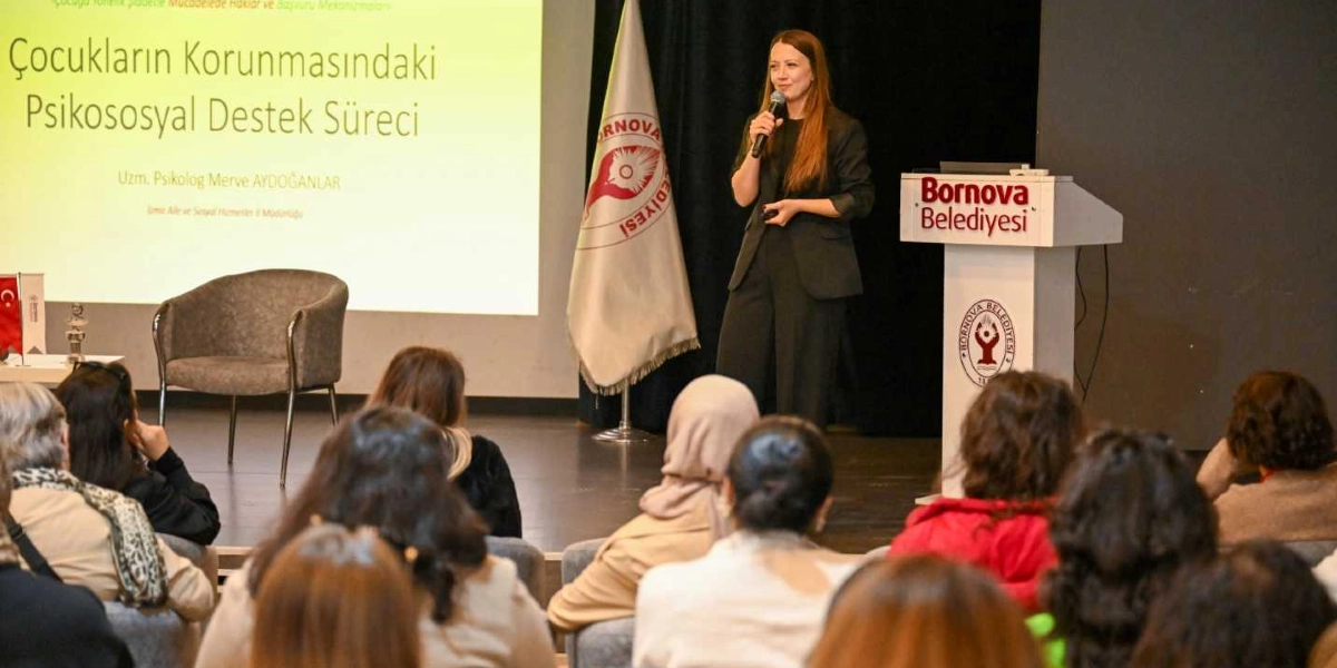 Uzman Psikolog Merve Aydoğanlar, Bornova Belediyesi tarafından düzenlenen bir etkinlikte kürsüde konuşma yapıyor. Sahnedeki projeksiyon ekranında 