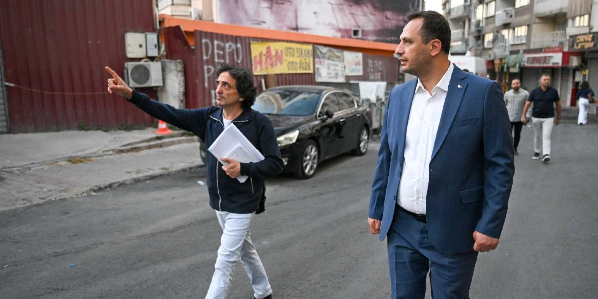 Koyu renkli bir ceket ve beyaz pantolon giymiş, elinde dosyalar tutan kıvırcık saçlı bir adam, yanında lacivert takım elbiseli bir başka adamla birlikte Bornova sokaklarında yürürken eliyle bir yeri işaret ediyor. Arka planda üzerinde 