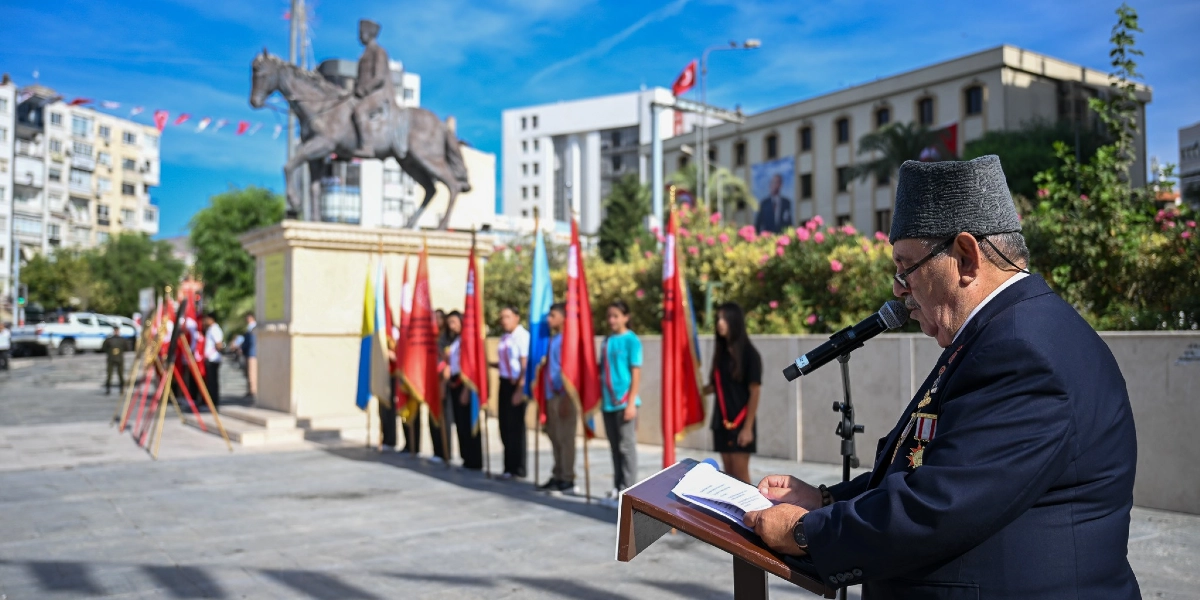 Bornova'da düzenlenen 19 Eylül Gaziler Günü töreninde, üzerinde madalyaları bulunan bir gazi kürsüde konuşma yapıyor. Arka planda Atatürk heykeli, ellerinde çeşitli bayraklar tutan gençler ve tören alanındaki katılımcılar yer alıyor.