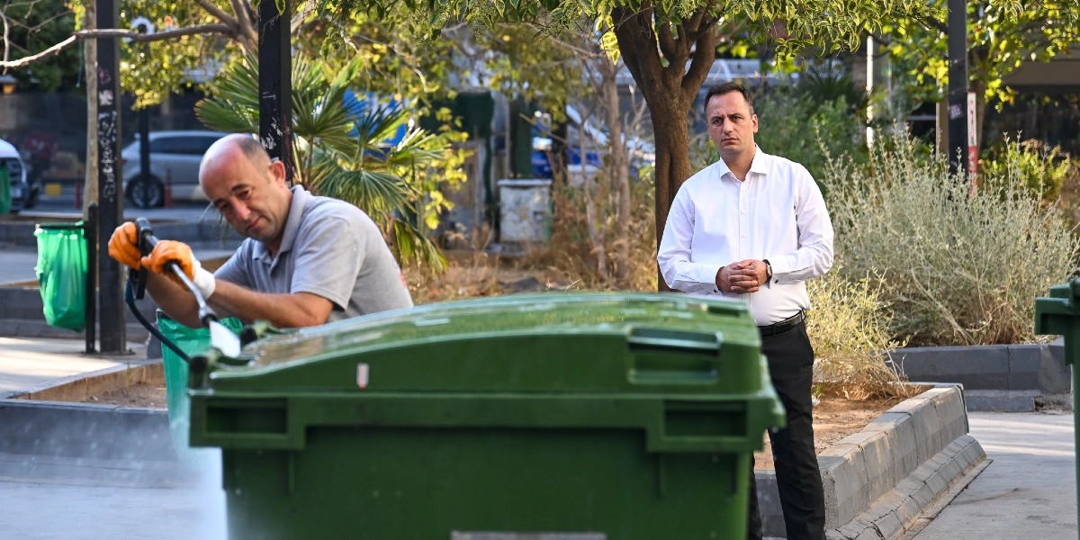 Bornova Belediye Başkanı Ömer Eşki, beyaz gömlek ve siyah pantolonuyla park benzeri bir alanda dururken, yanında bir belediye çalışanı basınçlı su makinesiyle yeşil bir çöp konteynerini temizliyor.
