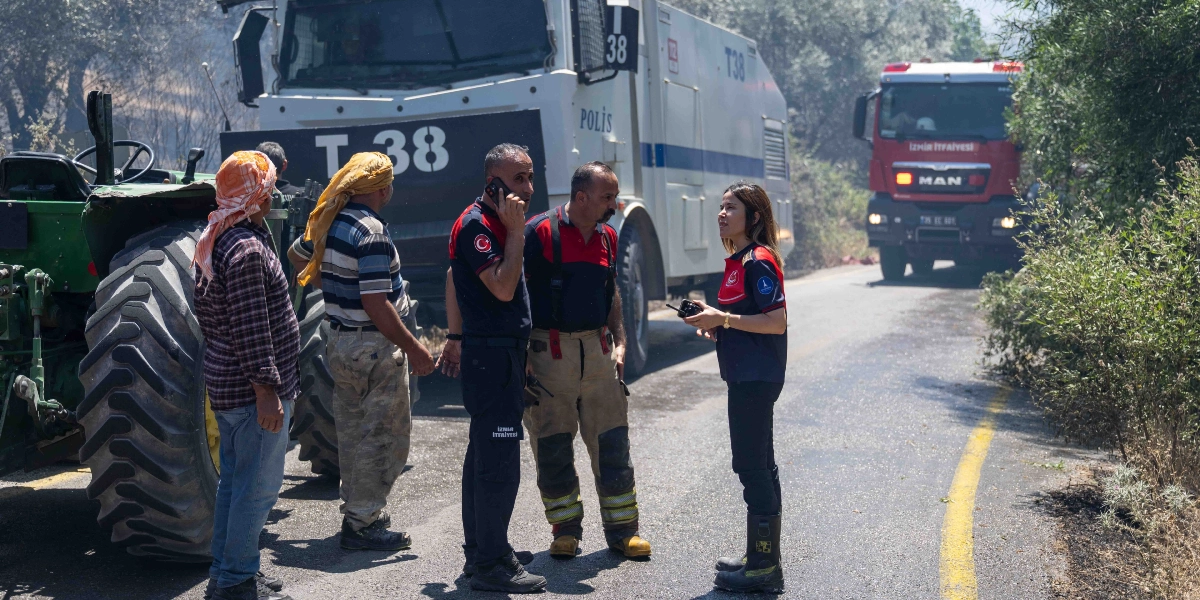 Bornova Homeros Vadisi'ndeki yangın bölgesinde, asfalt yol üzerinde duran iki sivil vatandaş, iki itfaiye personeli ve bir kadın yetkili koordinasyon sağlıyor. İtfaiye personelinin arkasında üzerinde T-38 yazan beyaz bir TOMA aracı, yolun ilerisinde ise İzmir İtfaiyesi'ne ait kırmızı bir itfaiye aracı bulunuyor. Ekip üyeleri telsiz ve telefonla iletişim kurarken, yan tarafta büyük bir traktör tekerleği görülüyor.