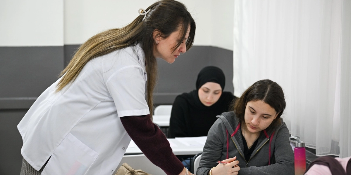 Beyaz önlüklü bir eğitmen, sınıfta ders çalışan iki öğrenciye rehberlik ediyor. Öğrencilerden biri gri fermuarlı bir sweatshirt giyerken, diğeri siyah bir başörtüsü takıyor. Eğitim ortamında öğrenciler masada oturarak not alıyor, eğitmen ise onlara doğru eğilerek destek veriyor.