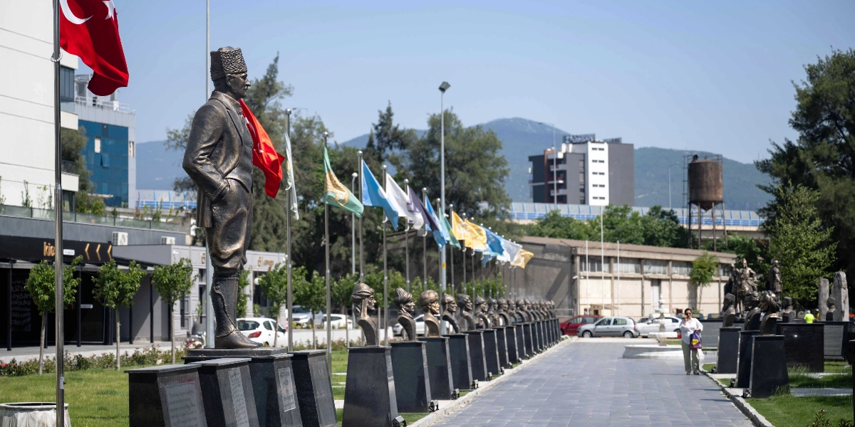 Bornova'da açık havada, bir yürüyüş yolu boyunca sıralanmış büstlerin ve en başta Mustafa Kemal Atatürk'ün tam boy heykelinin bulunduğu bir meydan. Heykellerin arkasında Türk bayrakları ve çeşitli renklerde flamalar dalgalanıyor. Arka planda modern binalar, ağaçlar ve dağ manzarası görülürken, yürüyüş yolunda bir kişi yürüyor.