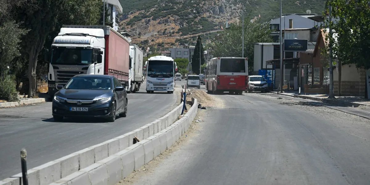 Pınar Caddesi'nde devam eden yol yenileme çalışmaları sırasında, ortada beton bariyerlerle bölünmüş asfalt yolda ilerleyen bir binek araç, tır, otobüs ve diğer araçların oluşturduğu trafik akışı görülüyor. Yolun sağ tarafında çalışma alanı ve binalar yer alırken, arka planda ağaçlıklı bir tepe manzarası bulunuyor.
