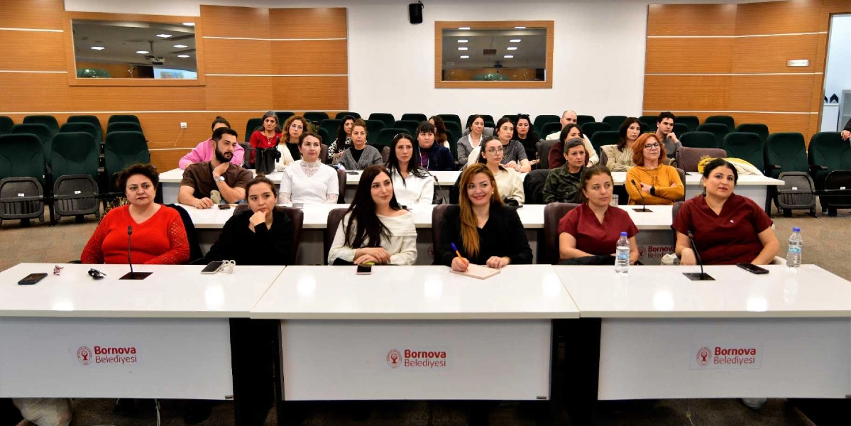 Bornova Belediyesi'nde düzenlenen bir toplantıda, beyaz masaların etrafında oturan ve çoğunluğu kadınlardan oluşan katılımcılar dinleyici sıralarında yer alıyor. Ön sırada oturan altı kişi, masaların üzerindeki mikrofonlar eşliğinde toplantıyı takip ediyor. Arka sıralarda ise daha kalabalık bir grup, ahşap panellerle kaplı duvarları olan konferans salonunda oturuyor. Masaların ön yüzünde Bornova Belediyesi logosu ve yazısı yer alıyor.