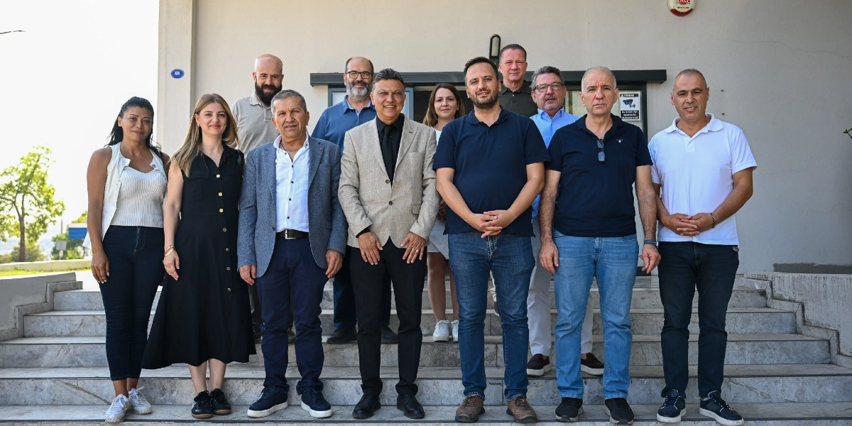 Bej renkli bir binanın girişindeki merdivenlerde yan yana duran, aralarında Bornova Belediye Başkanı Ömer Eşki'nin de bulunduğu on bir kişilik grup. Başkan Eşki, ön sırada, bej renkli ceket ve siyah gömlek giymiş şekilde merkezde yer alıyor. Grubun geri kalanı günlük kıyafetler içinde, farklı yaş ve tarzlarda kadın ve erkeklerden oluşuyor. Hepsi kameraya bakarak poz veriyor.