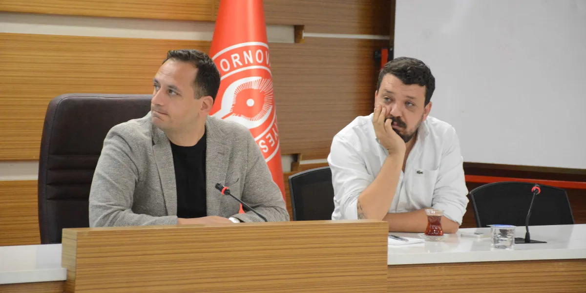 Toplantı masasında yan yana oturan iki erkekten soldaki gri ceketli ve siyah tişörtlü kişi sola doğru bakarken, sağdaki beyaz gömlekli kişi elini çenesine dayamış şekilde karşıya bakmaktadır. Masanın üzerinde mikrofonlar, bir bardak çay ve su bardağı bulunmaktadır. Arka planda Bornova Belediyesi'nin kırmızı renkli logosunun yer aldığı bir bayrak ve ahşap panelli bir duvar görünmektedir.
