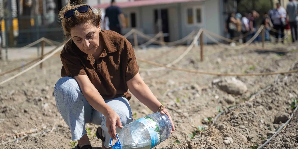 Kahverengi gömlekli ve açık mavi kot pantolonlu bir kadın, toprak bir alanda diz çökmüş, elindeki büyük plastik su bidonuyla yeni dikilmiş küçük bir bitkiyi suluyor. Arka planda iplerle çevrili bir bahçe alanı ve bulanık şekilde görünen insanlar yer alıyor.