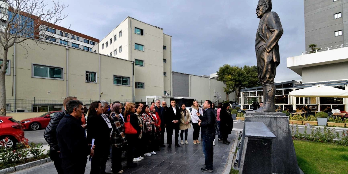 Açık havada, Bornova Muhtarlar Akademisi kapsamında bir araya gelen bir grup insan, Atatürk heykelinin önünde durarak bir konuşmacıyı dinliyor. Arka planda modern binalar, park halindeki kırmızı bir otomobil ve bir kafe alanı yer alıyor.