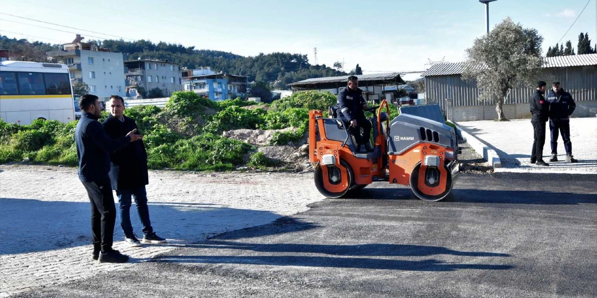 Bornova'da devam eden yol yapım çalışmaları kapsamında, asfalt serim alanında turuncu renkli bir silindir operatörü tarafından kullanılırken, iki kişi çalışmaları izleyerek sohbet ediyor. Arka planda ise iki görevli daha ayakta duruyor ve çevrede binalar ile yeşil alanlar bulunuyor.