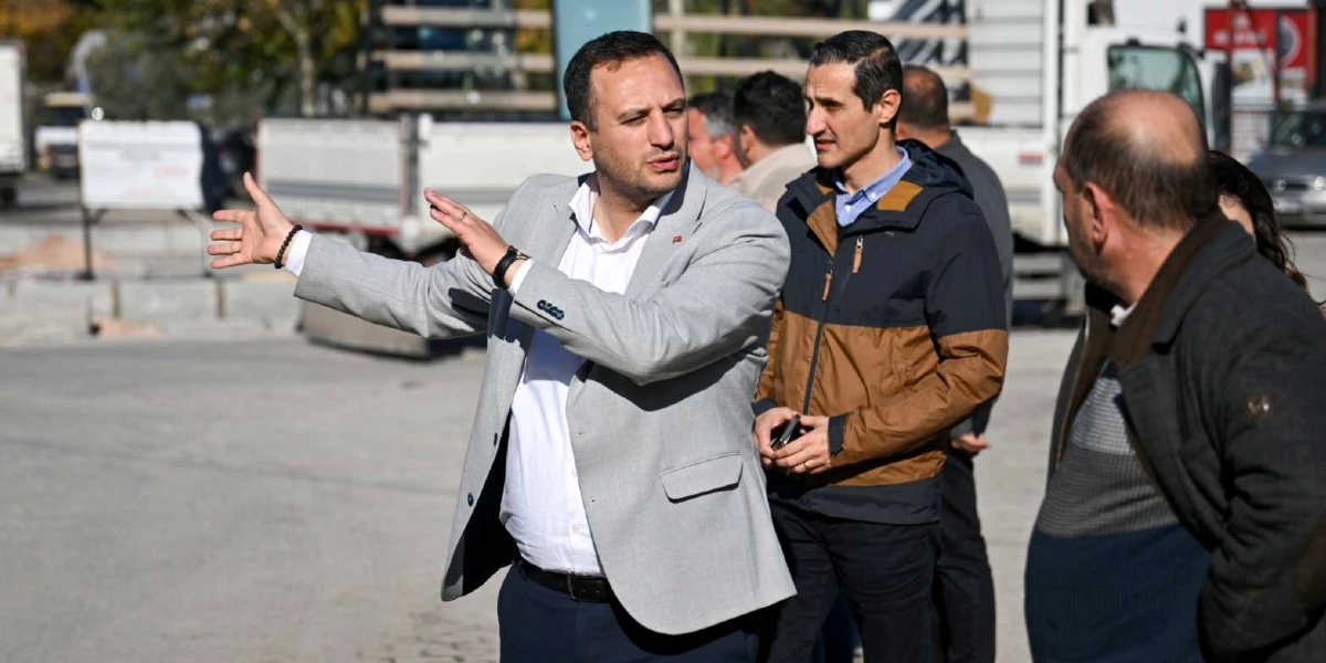 Açık renk ceketli bir yetkili, açık havada bir şantiye veya yol çalışması alanında el hareketleriyle çevresindekilere bilgi veriyor. Yanında kahverengi ve lacivert renkli bir mont giyen başka bir erkek ile sırtı dönük şekilde duran bir kişi daha bulunuyor. Arka planda ise inşaat araçları ve çalışma alanı görülüyor.