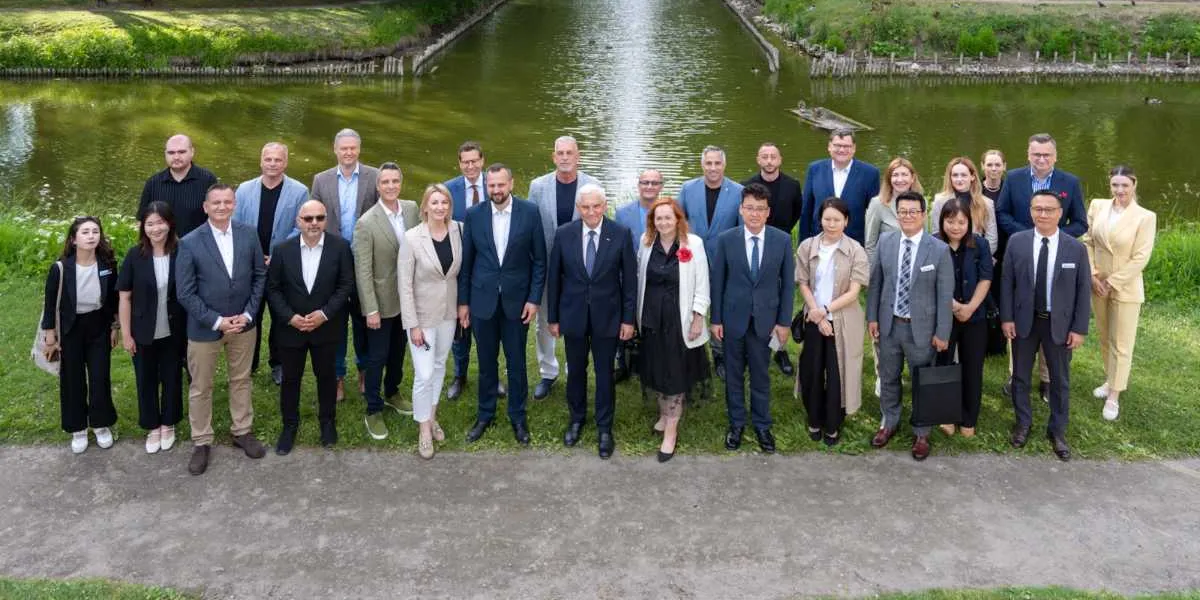 Yeşil bir park alanında, arkalarında bir kanalın bulunduğu, resmi kıyafetler giymiş yaklaşık 25 kişilik bir grup, yan yana dizilmiş şekilde kameraya gülümseyerek poz veriyor. Kadın ve erkeklerden oluşan bu çok uluslu heyet, iş birliği ve diplomatik temasları simgeleyen bir etkinlik kapsamında bir arada bulunuyor.