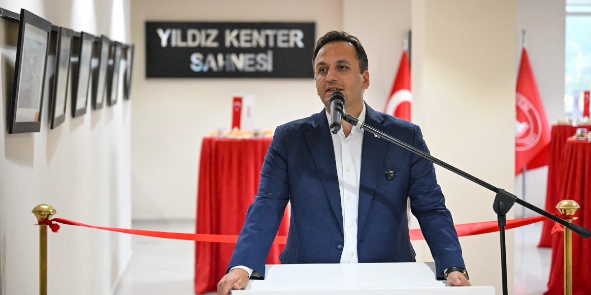 Yıldız Kenter Sahnesi yazılı tabelanın önünde, beyaz gömlek ve lacivert ceket giymiş bir erkek, kürsüde mikrofon başında konuşma yapıyor. Arka planda kırmızı örtülü masalar ve Türk bayrakları yer alıyor, sol taraftaki duvarda ise çerçeveli fotoğraflar asılı bulunuyor.