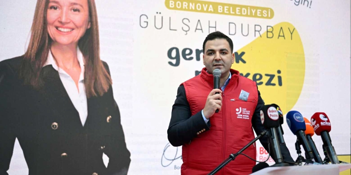 Üzerinde CHP Gençlik Kolları Genel Başkanı Cem Aydın yazılı kırmızı bir yelek giyen bir erkek, mikrofon tutarak konuşma yapıyor. Arka planda Bornova Belediyesi'ne ait, üzerinde Gülşah Durbay isminin ve gençlik merkezi ibaresinin yer aldığı büyük bir afiş bulunuyor. Afişin sol tarafında Gülşah Durbay'ın bir fotoğrafı yer alıyor, konuşmacının önünde ise çeşitli basın kuruluşlarına ait mikrofonlar duruyor.