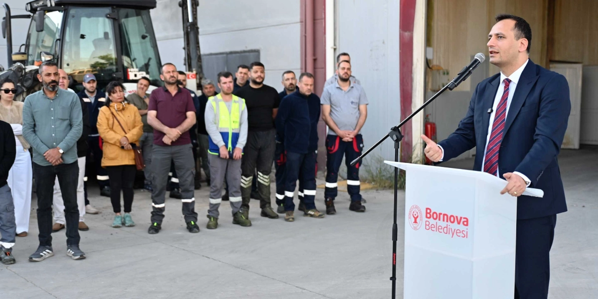 Lacivert takım elbiseli ve çizgili kravatlı bir erkek, üzerinde Bornova Belediyesi logosu bulunan bir kürsüde mikrofonla konuşma yapıyor. Konuşmacının karşısında, bir şantiye veya depo alanında duran, farklı kıyafetler giymiş bir grup çalışan onu dinliyor. Arka planda büyük bir iş makinesinin ön kısmı ve depo binasının girişi görülüyor.