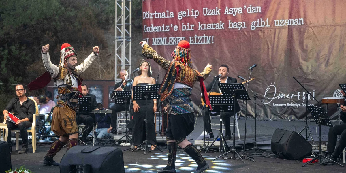 Açık havada kurulan bir sahnede, geleneksel zeybek kıyafetleri giymiş iki erkek dansçı performans sergiliyor. Dansçıların arkasında, Nazım Hikmet’in 