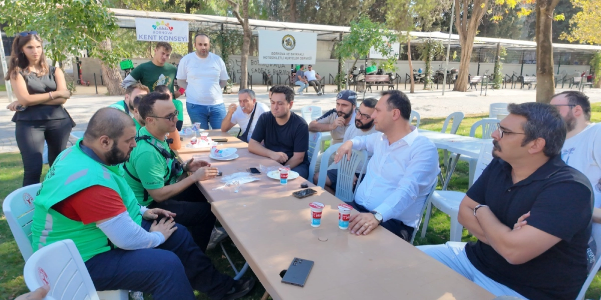 Açık havada, park benzeri bir alanda uzun bir masanın etrafında toplanmış bir grup insan. Masada oturanlar arasında üzerinde parlak yeşil yelekler olan moto kuryeler ve beyaz gömlekli bir yetkili bulunuyor. Arka planda 