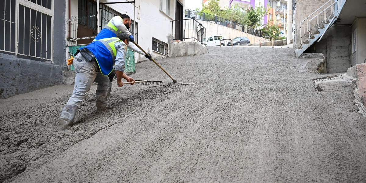 İki belediye işçisi, Bornova'da eğimli bir sokakta yeni dökülen taze betonu tırmık ve kürek yardımıyla düzeltiyor. İşçiler üzerinde reflektörlü yelekler ve koruyucu çizmeler bulunuyor.