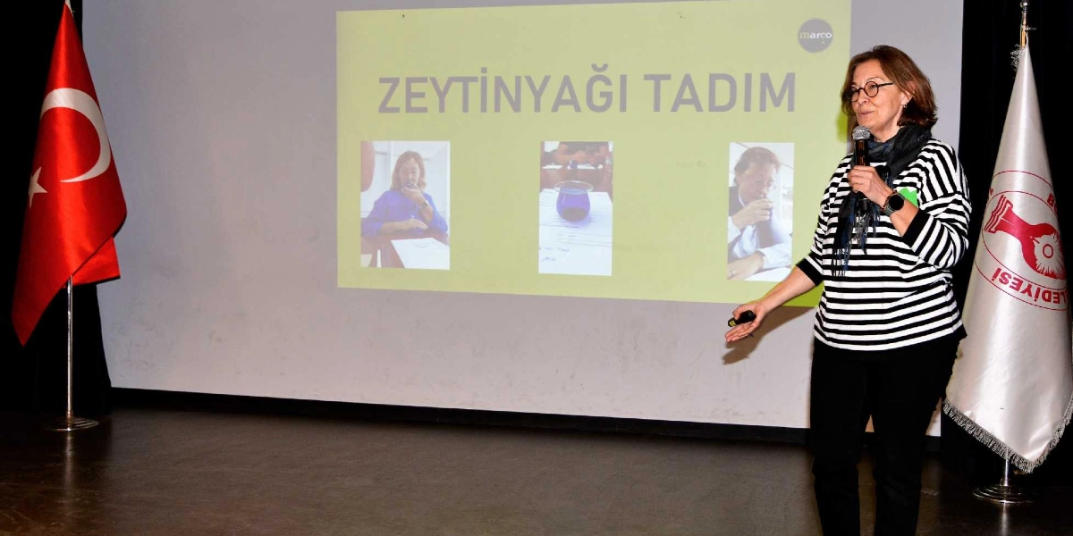 Siyah beyaz çizgili bluz giyen gözlüklü bir kadın, elinde mikrofonla sahnede konuşma yapıyor. Arkasındaki projeksiyon perdesinde 