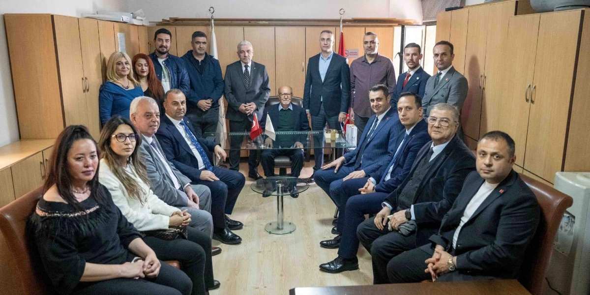 Ofis ortamında bir araya gelmiş, çoğunluğu takım elbiseli erkeklerden ve birkaç kadından oluşan kalabalık bir grup. Arka planda ahşap dolaplar yer alırken, grubun merkezinde masada oturan bir kişi ve çevresinde ayakta veya oturarak poz veren katılımcılar bulunuyor. Masanın üzerinde küçük bir Türk bayrağı yer alıyor.
