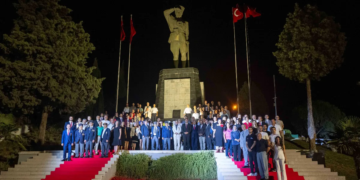 Gece vakti aydınlatılmış bir Atatürk anıtının önündeki geniş merdivenlerde, resmi kıyafetli ve sivil giyimli kalabalık bir grup, 30 Ağustos Zafer Bayramı kutlamaları kapsamında hatıra fotoğrafı çektiriyor. Merdivenlerin ortasında kırmızı bir halı serili olup, anıtın iki yanında Türk bayrakları dalgalanıyor.