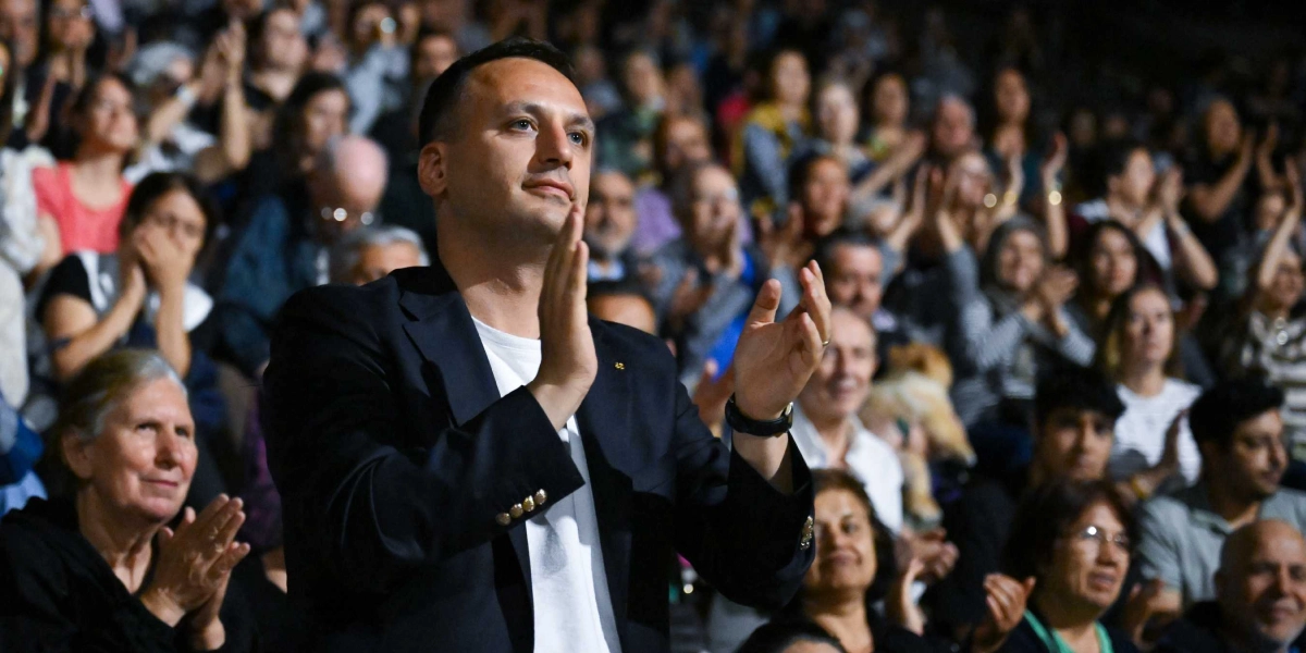 Koyu renk ceket ve beyaz tişört giymiş bir erkek, kalabalık bir izleyici kitlesinin önünde ayakta durarak elleriyle alkışlıyor. Arka planda yer alan çok sayıda kişi de aynı şekilde alkışlayarak etkinliğe katılım gösteriyor.