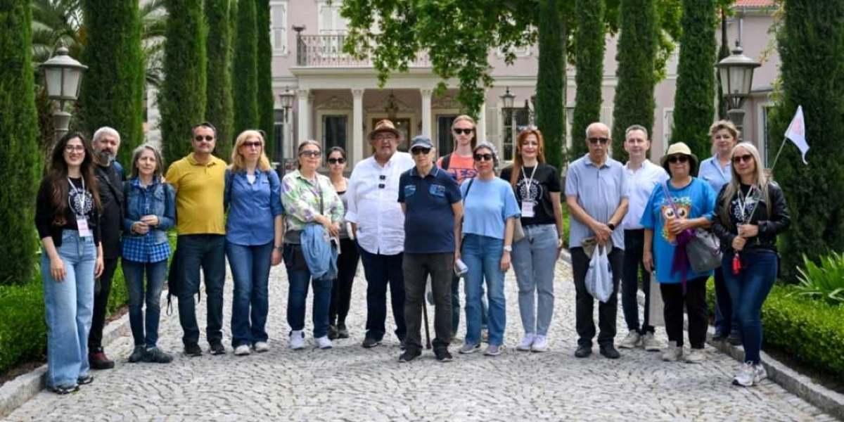 Tarihi bir köşkün önündeki taş yolda yan yana duran, farklı yaş gruplarından oluşan yaklaşık yirmi kişilik bir grup. Arka planda uzun selvi ağaçları ve klasik mimariye sahip, açık renkli, balkonlu bir yapı yer alıyor. Grup üyeleri günlük kıyafetler içinde, bazıları boyunlarında etkinlik kartlarıyla kameraya gülümseyerek poz veriyor.