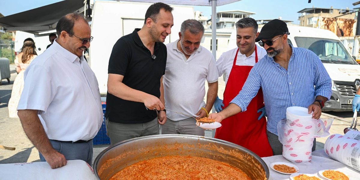 Açık havada kurulan bir stantta, büyük bir kazanda pişirilen menemenin başında duran beş erkek, gülümseyerek yemek dağıtımı yapıyor. Siyah tişörtlü bir kişi kepçeyle kazandan aldığı menemeni, yanında duran ve mavi çizgili gömlek giyen bir kişiye uzatıyor. Kırmızı önlük takmış bir kişi ise servis sürecine eşlik ediyor. Masanın üzerinde üst üste dizilmiş plastik tabaklar ve porsiyonlanmış yemek tabakları bulunuyor.