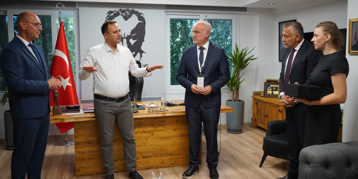 Modern bir ofis ortamında, ahşap bir çalışma masasının önünde duran beş kişilik bir heyet yer alıyor. Beyaz gömlekli bir kişi elleriyle açıklama yaparken, yanında lacivert takım elbiseli iki erkek ve siyah elbiseli bir kadın bulunuyor. Masanın üzerinde 