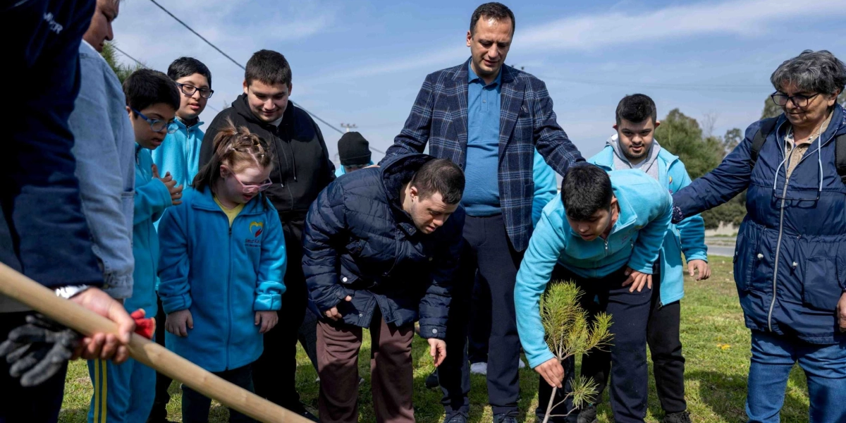 Açık havada, güneşli bir günde, özel gereksinimli gençlerden oluşan bir grup, yanlarındaki yetişkinlerle birlikte fidan dikiyor. Mavi ceketli gençler toprağa yerleştirilen küçük bir çam fidanına dikkatle bakarken, gruptaki bir genç fidanı eliyle destekliyor. Çevredeki katılımcılar merakla süreci izliyor.