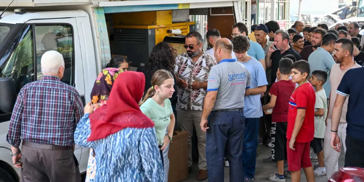 Bornova Belediyesi tarafından düzenlenen bir etkinlikte, beyaz bir aracın yanına kurulan stanttan yemek veya ikram alan kalabalık bir grup insan görülüyor. Ön planda başörtülü kadınlar ve çocuklar yer alırken, arka planda farklı yaş gruplarından erkekler sıranın ilerlemesini bekliyor. Görevli bir kişi, üzerinde 