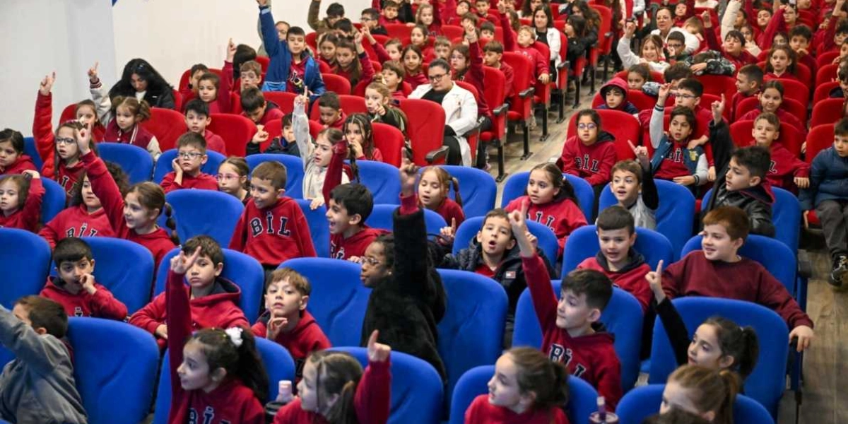 Konferans salonunda oturan, üzerinde kırmızı sweatshirtler bulunan ilkokul ve ortaokul öğrencileri, bir soruya cevap vermek için heyecanla ellerini kaldırmış durumdalar. Mavi koltuklarda oturan öğrenciler, dikkatlerini ön tarafa yöneltmiş şekilde aktif bir katılım sergiliyor.