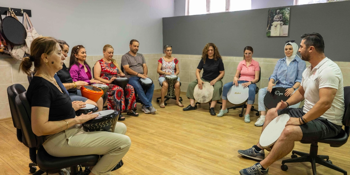 Bir eğitmen eşliğinde çember şeklinde oturmuş, ellerinde darbuka ve benzeri vurmalı çalgılar tutan yetişkinlerden oluşan bir grup müzik atölyesi çalışması yapıyor.