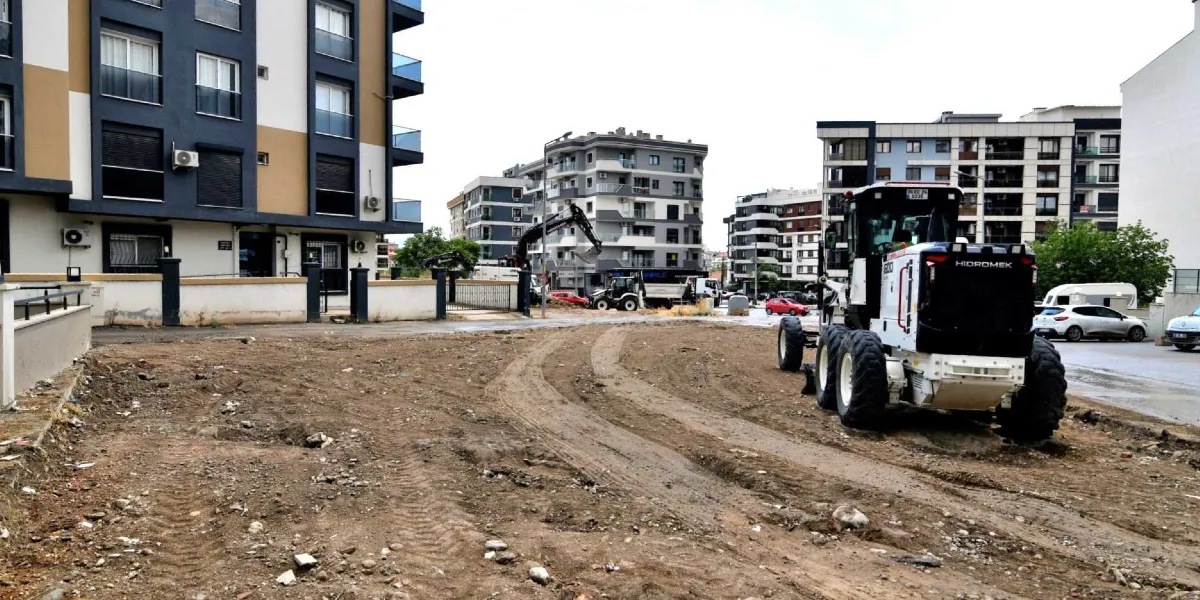 Şehir içindeki boş bir arazide çalışma yapan beyaz renkli bir Hidromek marka greyder ve arka planda kazı yapan bir iş makinesi görülüyor. Çevrede çok katlı apartmanlar ve park halindeki araçlar yer alıyor.