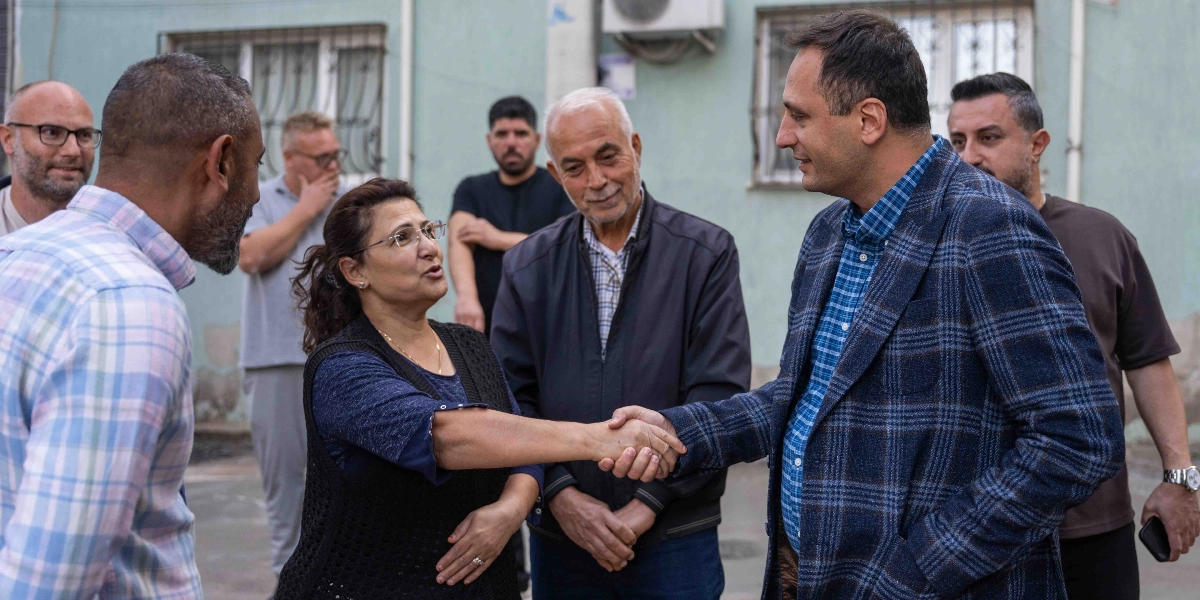 Mavi ekose ceketli bir erkek, sokakta bir kadınla el sıkışırken çevresinde onları izleyen bir grup erkek bulunuyor. Kadın, lacivert bir bluz ve üzerine siyah yelek giymiş, gözlüklü bir şekilde karşısındaki kişiyle konuşuyor. Arka planda ise açık yeşil renkli bir bina duvarı yer alıyor.